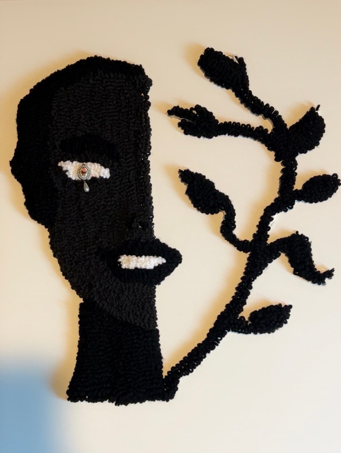 Black textile art of a face profile with a jeweled eye and leafy branch.